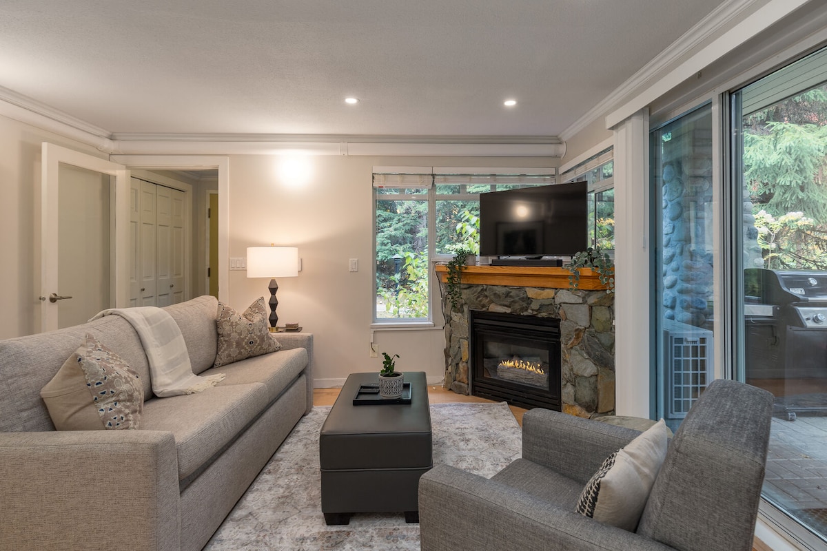A living room is presented with a cozy gas fireplace and a television mounted above. A gray sofa and an armchair are arranged around a central coffee table, with large windows providing natural light and views of greenery outside.