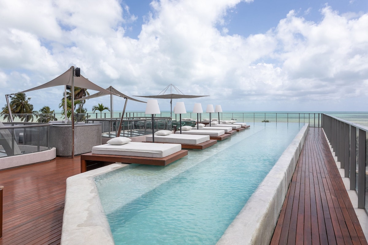 A rooftop pool is featured with a sleek design, surrounded by wooden decking and lounge chairs. Umbrellas provide shade, while the ocean stretches out in the background under a cloudy sky. Lush palm trees can be seen in the distance, enhancing the tranquil setting.