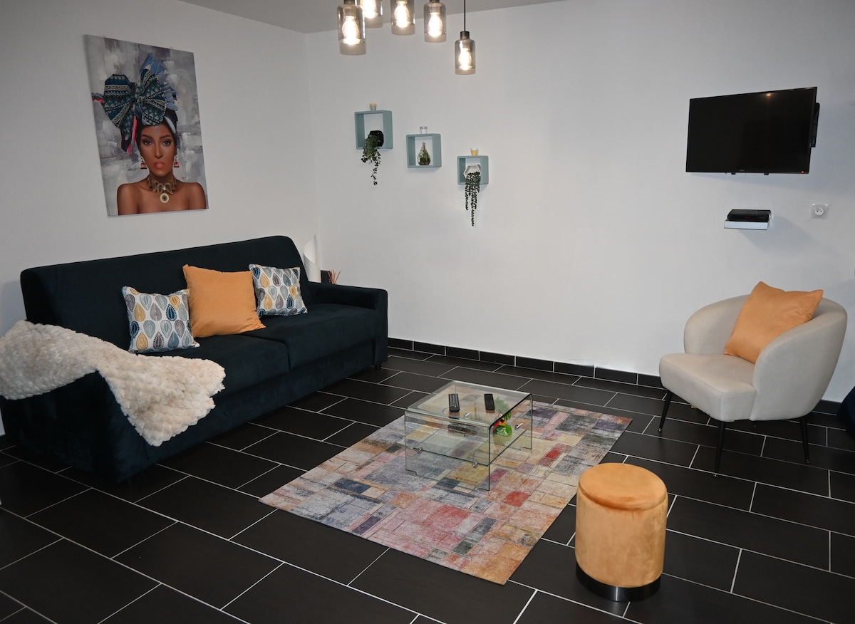 A modern living area features a dark green sofa adorned with decorative pillows, complemented by an accent chair. A glass coffee table sits on a colorful area rug, and wall-mounted frames display art. A flat-screen TV is mounted on the wall, enhancing the space.