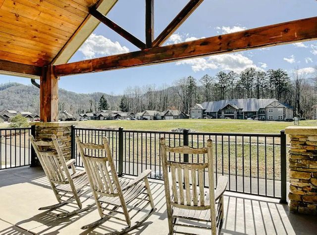 A spacious covered deck features rustic rocking chairs, providing a comfortable seating area with views of the surrounding landscape. Lush trees and distant mountain peaks are visible in the background, enhancing the serene atmosphere of the outdoor space.