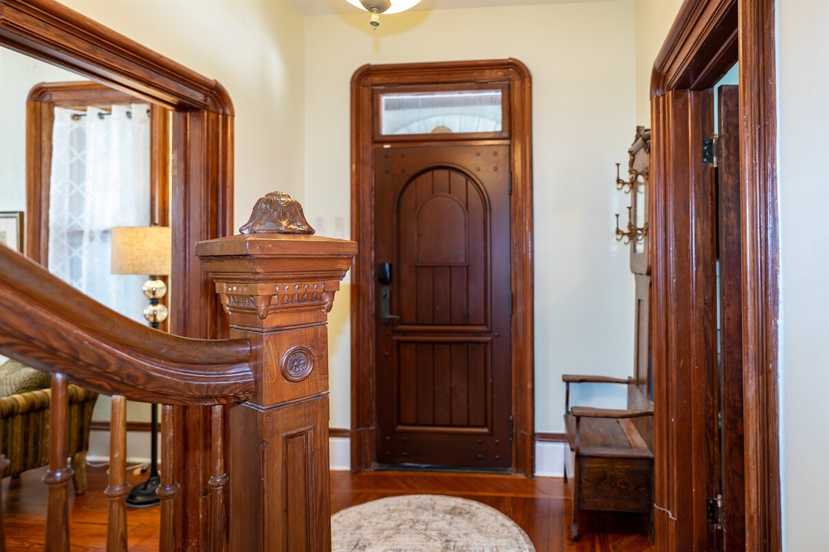 A welcoming entryway showcases original architectural details, including rich wooden trim and a carved banister. The solid front door, framed by a decorative archway, opens to the inviting interior. A small bench and coat rack enhance the functional space, while a soft area rug adds warmth.