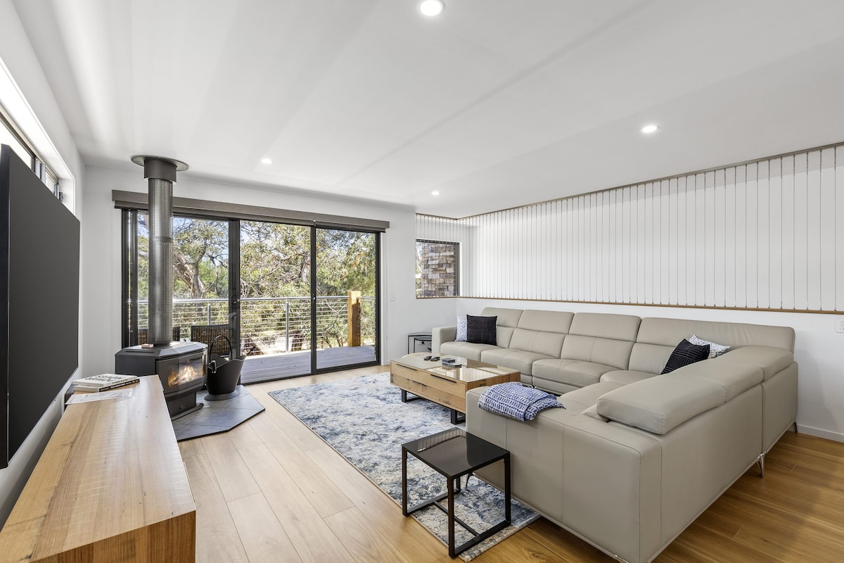 A modern living area features a light-colored sectional sofa, a wooden coffee table, and a flat-screen TV mounted on the wall. Large glass doors provide views of the outdoors and let in natural light. A wood fire is positioned beside the seating area.