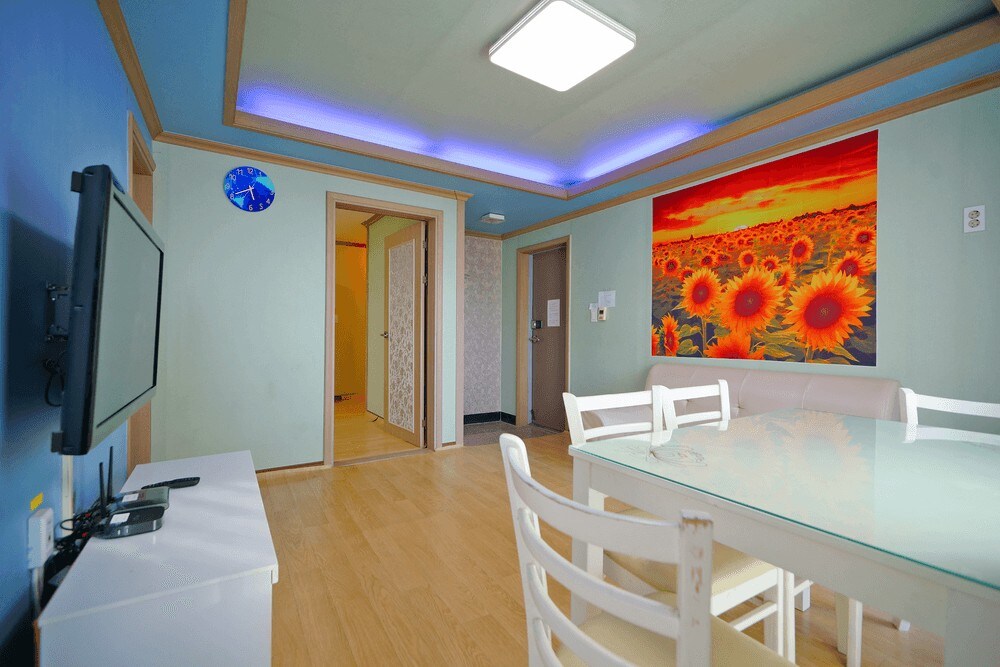 An open living area features a glass dining table surrounded by white chairs. A large artwork of sunflowers adorns the wall, complemented by a blue accent wall. A television is mounted opposite the dining space, enhancing the room's functionality.