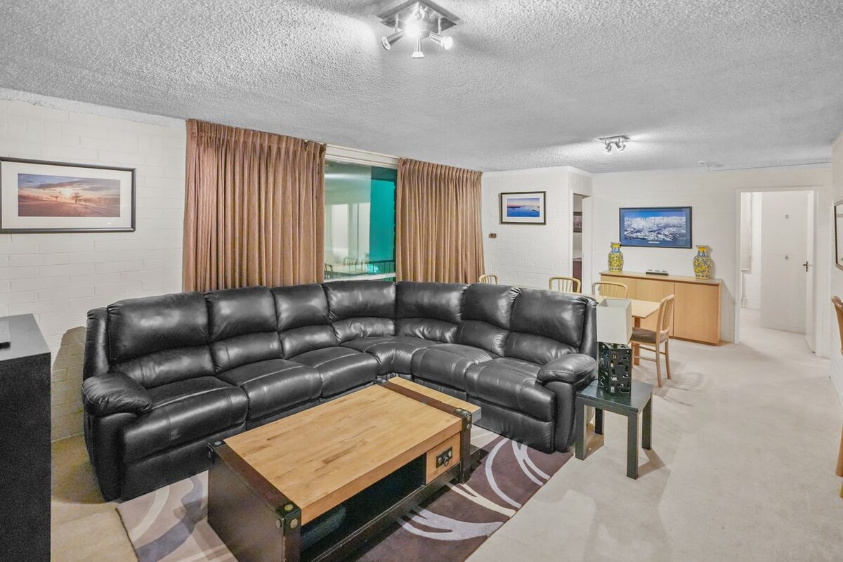 A spacious living area features a large black leather sectional sofa arranged around a wooden coffee table. Natural light filters in through expansive windows, while darker curtains offer privacy. A dining area with a table and chairs is seen in the background, complementing the cozy atmosphere.