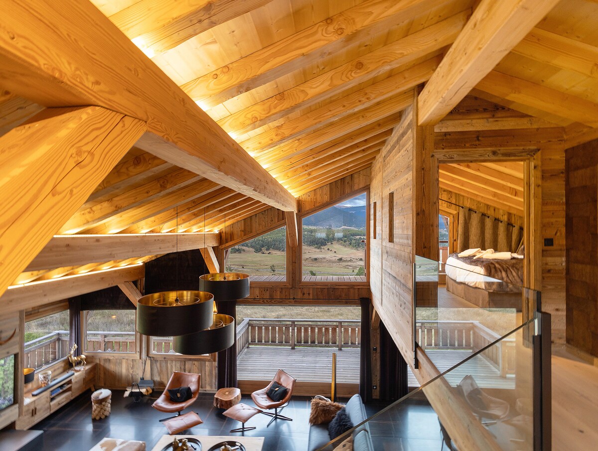 The open-concept living space features high wooden ceilings and expansive windows that frame views of the surrounding landscape. Comfortable seating areas are arranged around a central table, creating an inviting atmosphere. A partial view of a lofted sleeping area adds a sense of spaciousness.