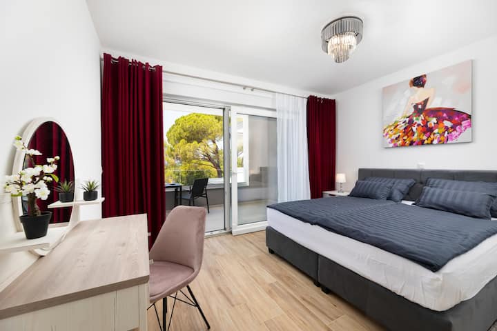 Prostrana spavaća soba s velikim krevetom i crvenim zavjesama te balkonom — idealna za opuštanje i jutarnju kavu na svježem zraku.
