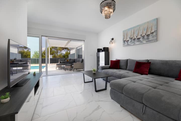 Svijetli i prostrani dnevni boravak s velikom sivom kutnom garniturom, crvenim i crnim jastucima te velikim kliznim vratima koja vode prema bazenu.