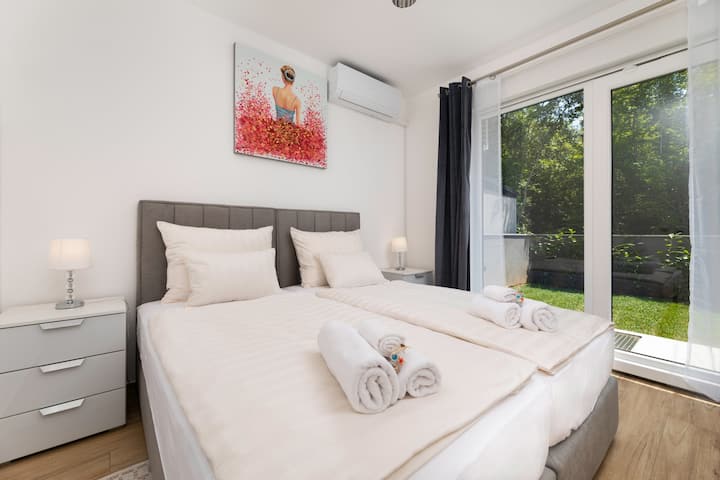  Svijetli kut sobe s velikim prozorom i pogledom na zelenilo — mirno mjesto za odmor.

