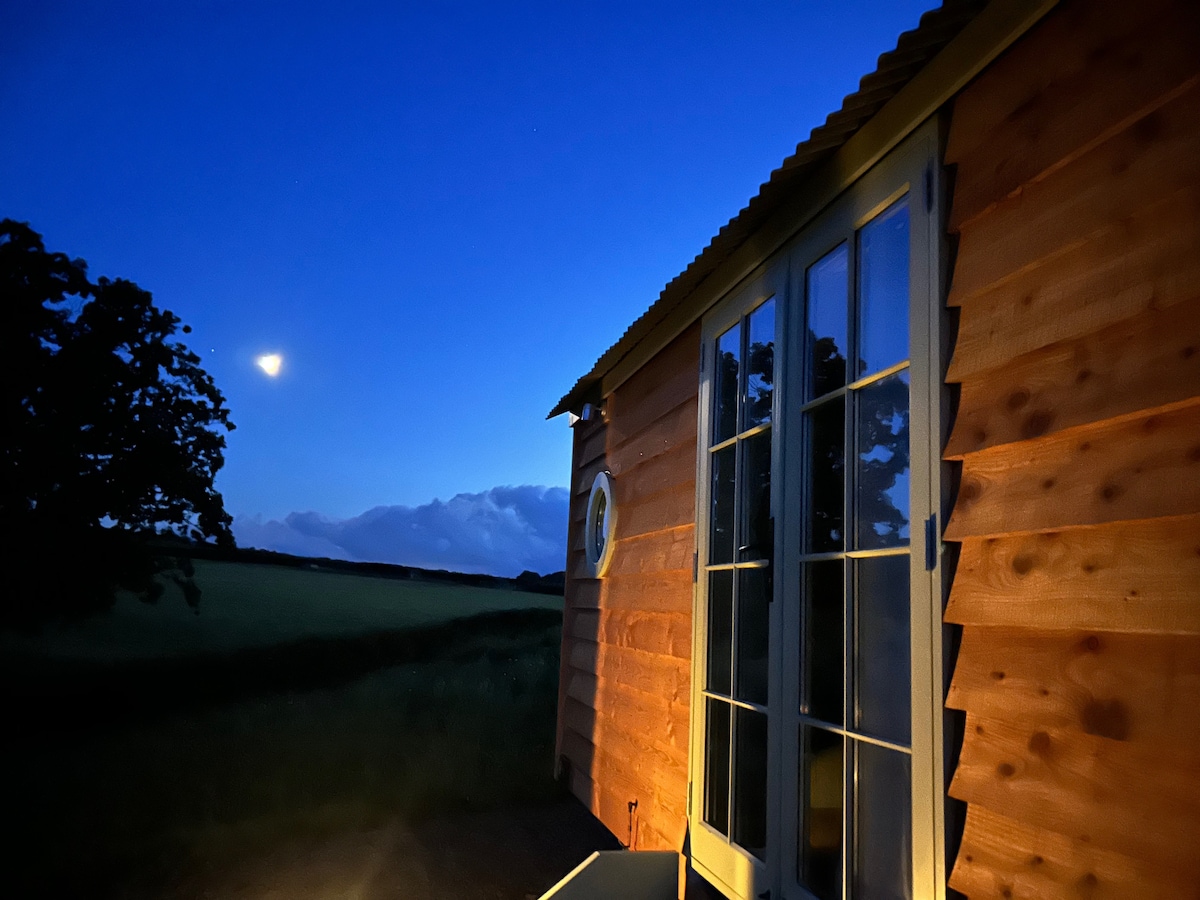 The exterior of the Shepherds hut is illuminated softly by warm light, showcasing rustic wooden cladding. A crescent moon can be seen above, with the evening sky transitioning into darker shades. The surrounding landscape features open fields, creating a serene and peaceful atmosphere.