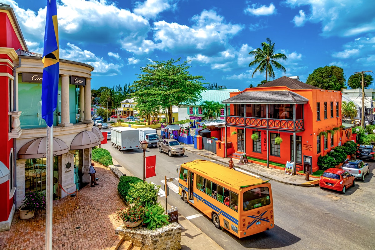 A vibrant streetscape features colorful buildings, including a red structure with a balcony and a yellow bus passing by. Lush trees provide shade along the road, which is lined with cars and trucks. The sky is bright with scattered clouds, creating a lively atmosphere.
