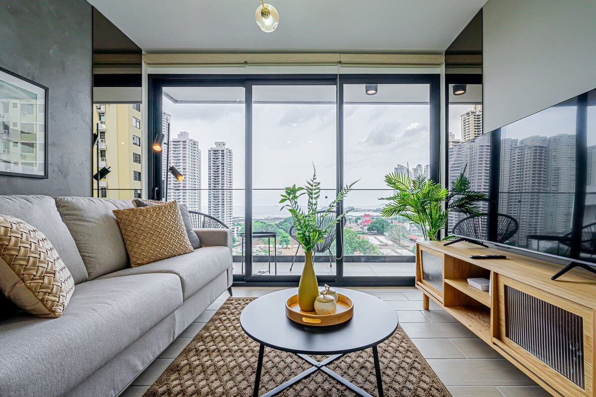 A spacious living area is designed with a light-colored sofa and a dark coffee table. Large glass doors reveal a balcony with views of the city skyline. A decorative plant and a vase are placed on the table, complementing the contemporary decor.