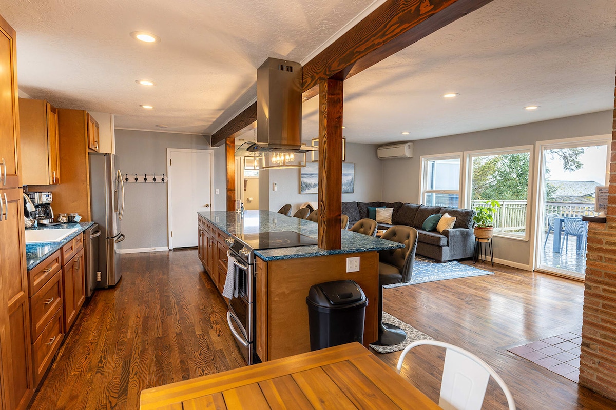 The modern kitchen features stainless steel appliances and a large island with stone countertops, providing ample space for meal preparation. A neighboring dining area with a wooden table invites communal dining, while the living area is visible through expansive windows that showcase natural light and views.
