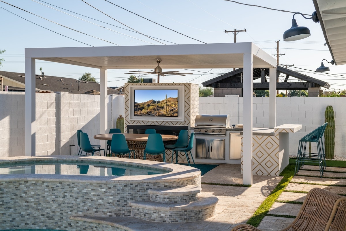 An outdoor dining area is featured under a shaded pergola, equipped with a barbecue grill and a television. A round table with teal chairs is positioned nearby, and a sparkling pool reflects the surrounding landscape, creating a welcoming space for relaxation and entertainment.