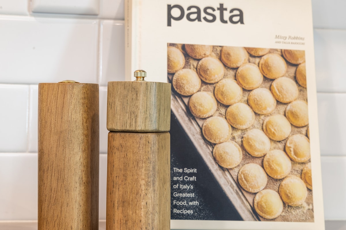 Two wooden pepper mills stand side by side in front of a cookbook titled 'Pasta.' The book features an appealing image of pasta in a rustic setting, emphasizing Italian culinary traditions, while the minimalist kitchen background showcases white tiled walls.