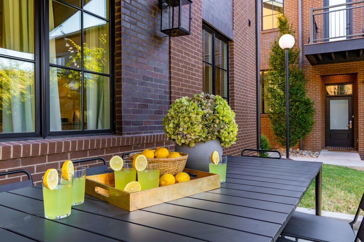 An outdoor patio area features a dark metal dining table set for refreshments. Glasses filled with bright green beverages sit alongside a wooden tray with lemons and a basket of oranges. Lush greenery is visible in the background against a red-brick building facade.