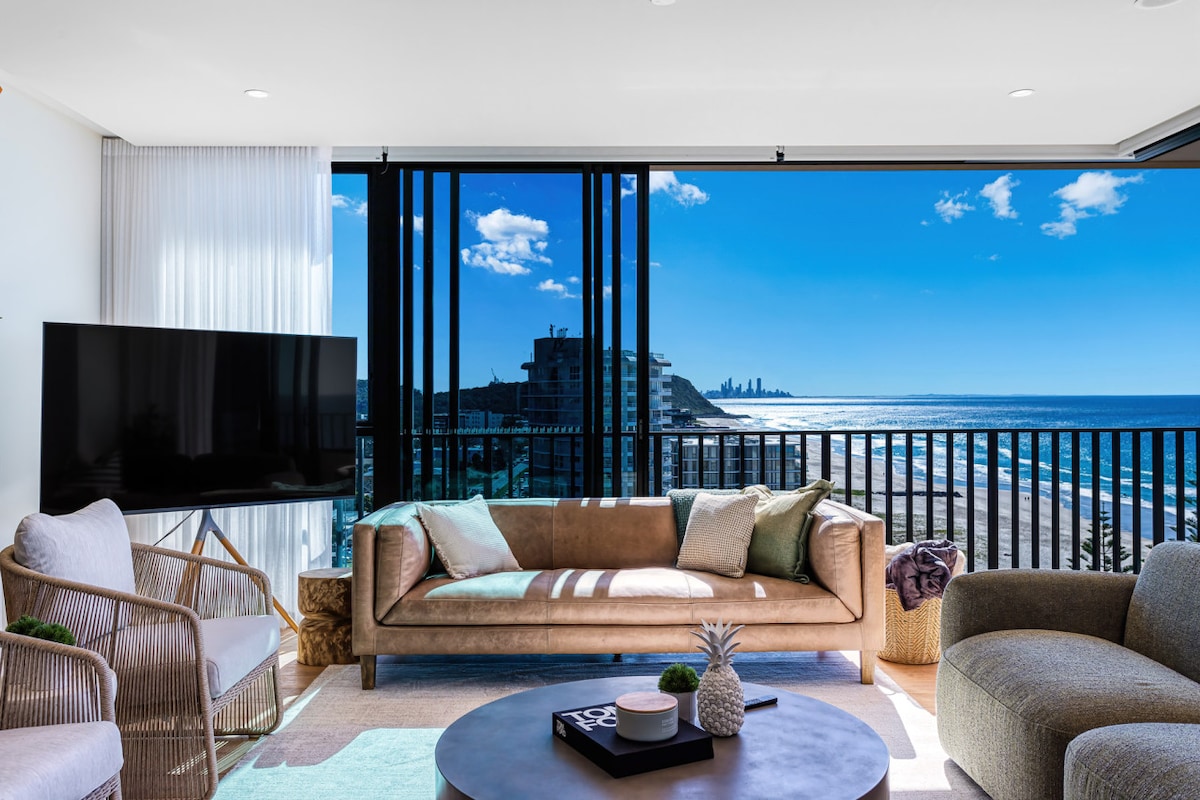 A bright living area features contemporary furnishings, including a plush sofa and comfortable chairs. Floor-to-ceiling windows offer sweeping ocean views and abundant natural light, while a large smart TV is positioned nearby. A coffee table sits centrally, adding functionality to the space.