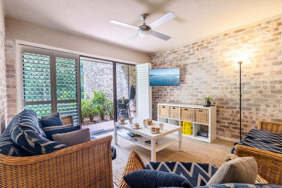 A cozy living area is highlighted with wicker furniture and navy accents. A coffee table is set in the center, and light from the window illuminates the brick walls. A Smart TV is mounted on a nearby wall, with greenery visible through the glass doors.