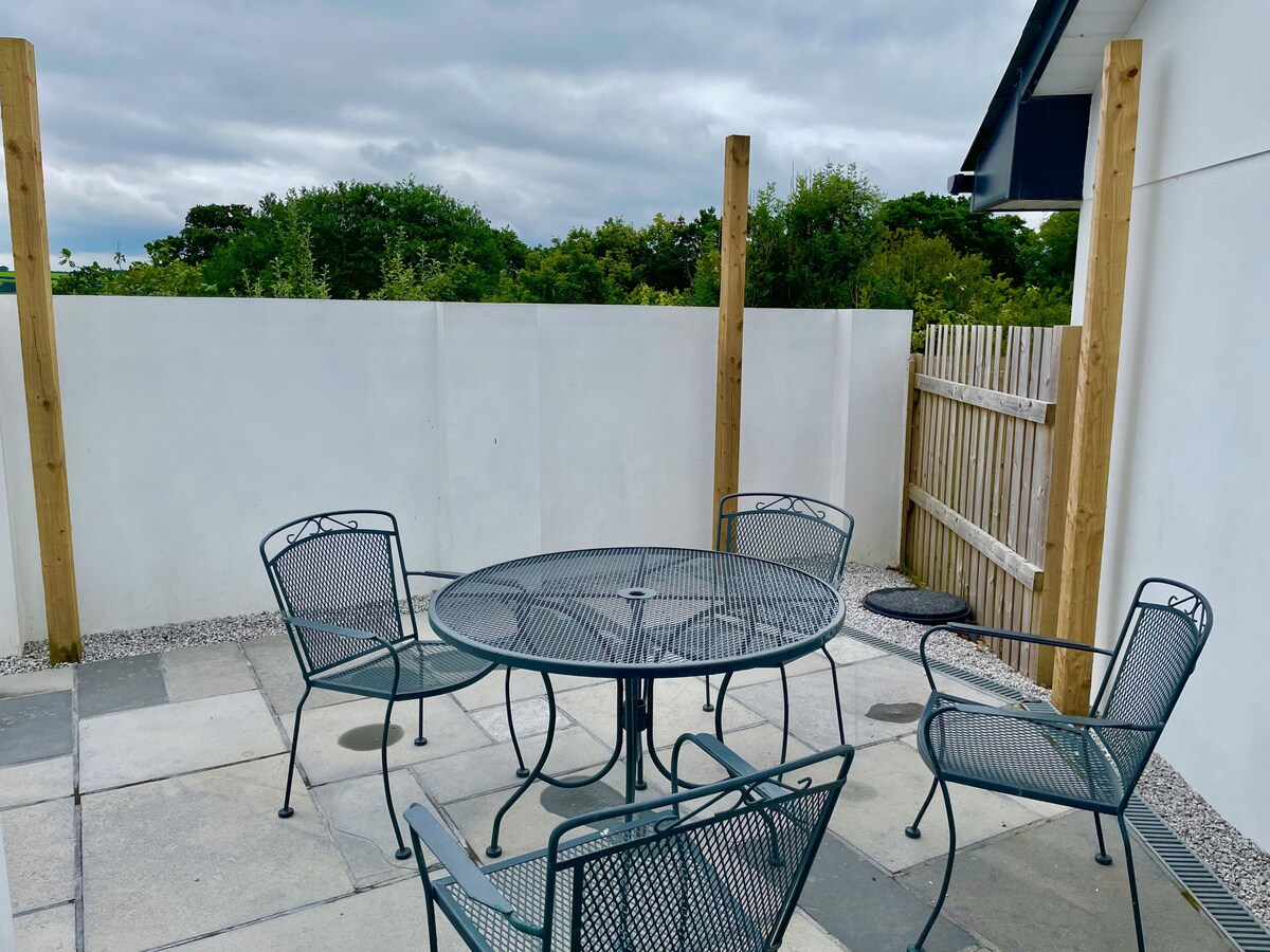 A patio area features a round table surrounded by four metal chairs. The space is enclosed by a low white wall, with greenery visible in the background. The stone flooring provides a neutral base, enhancing the inviting setting.