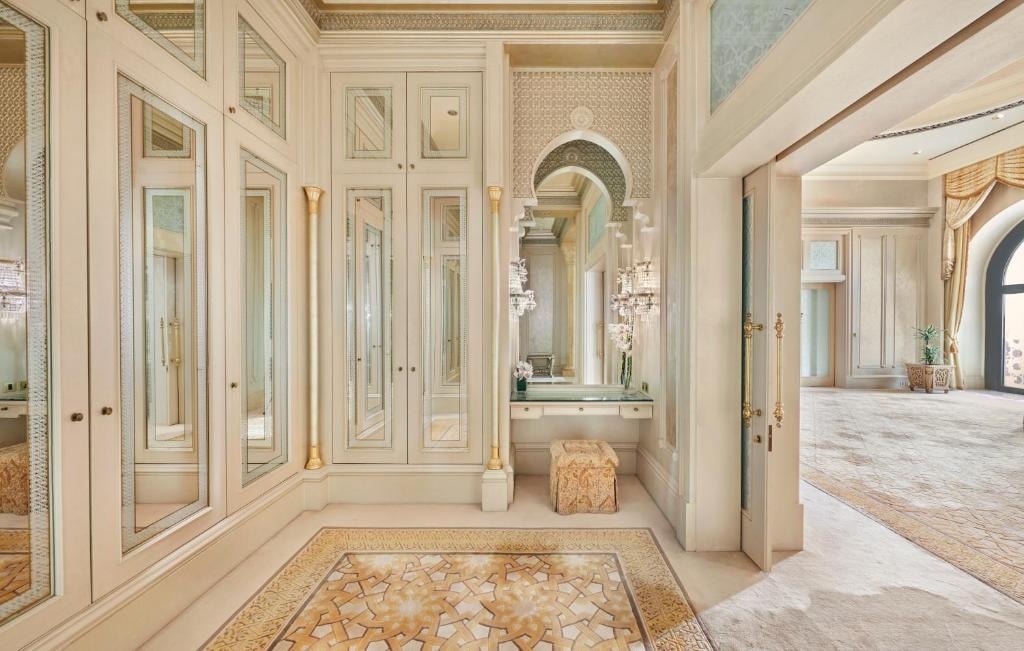 A spacious dressing area features mirrored wardrobes and an elegant vanity with soft lighting. Intricate tile work adorns the floor, creating a refined aesthetic. The room opens to a generous area, with soft textures and warm colors contributing to a sophisticated ambiance.