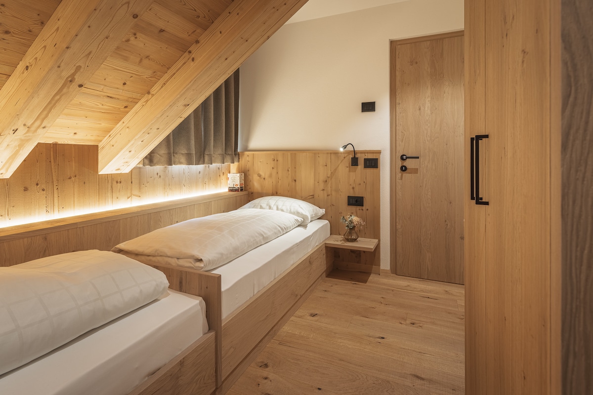 The bedroom features two single beds positioned against a wooden wall with soft lighting. A nightstand is placed beside one bed, decorated with a small plant. The slanted wooden ceiling adds warmth to the space, complemented by neutral tones and a door leading to the hallway.