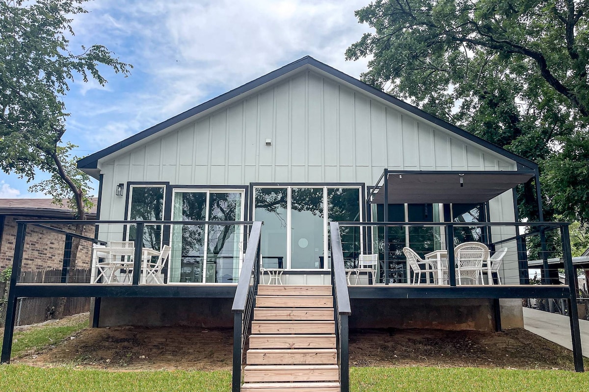 The exterior of a modern single-story property is displayed, featuring large glass windows and a spacious deck. A ramp leads up to the entryway, while outdoor seating is arranged on the deck, offering a comfortable space for relaxation. Surrounding trees provide shade.