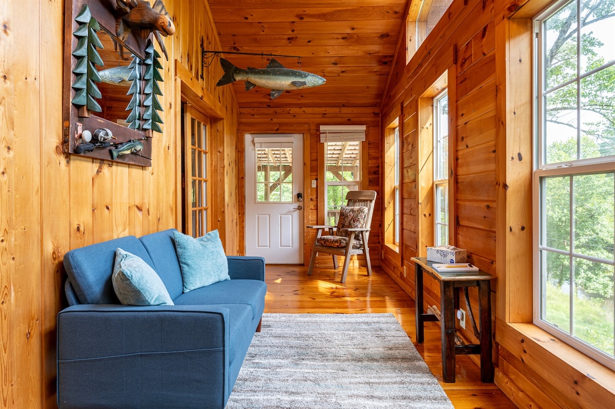 A sunlit sitting area features warm wooden walls and large windows, creating an inviting space. A blue couch with decorative cushions is positioned on a light rug, complemented by a rustic side table. Natural elements are showcased through decorative artwork, including a fish and wooden trees.