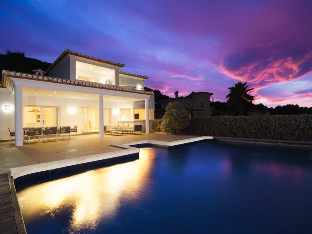 The exterior view of the villa showcases a modern design with outdoor seating arranged around a private pool. The pool reflects the vibrant colors of the sunset sky, creating a serene atmosphere. The two-story structure features large windows, blending interior and exterior spaces.