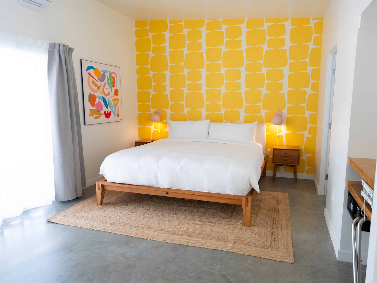 The room features a spacious bed with crisp white linens, complemented by a vibrant yellow accent wall. A colorful artwork hangs above a wooden nightstand on one side, while soft lighting is provided by lamps. Natural light enters through a window covered by light gray curtains.
