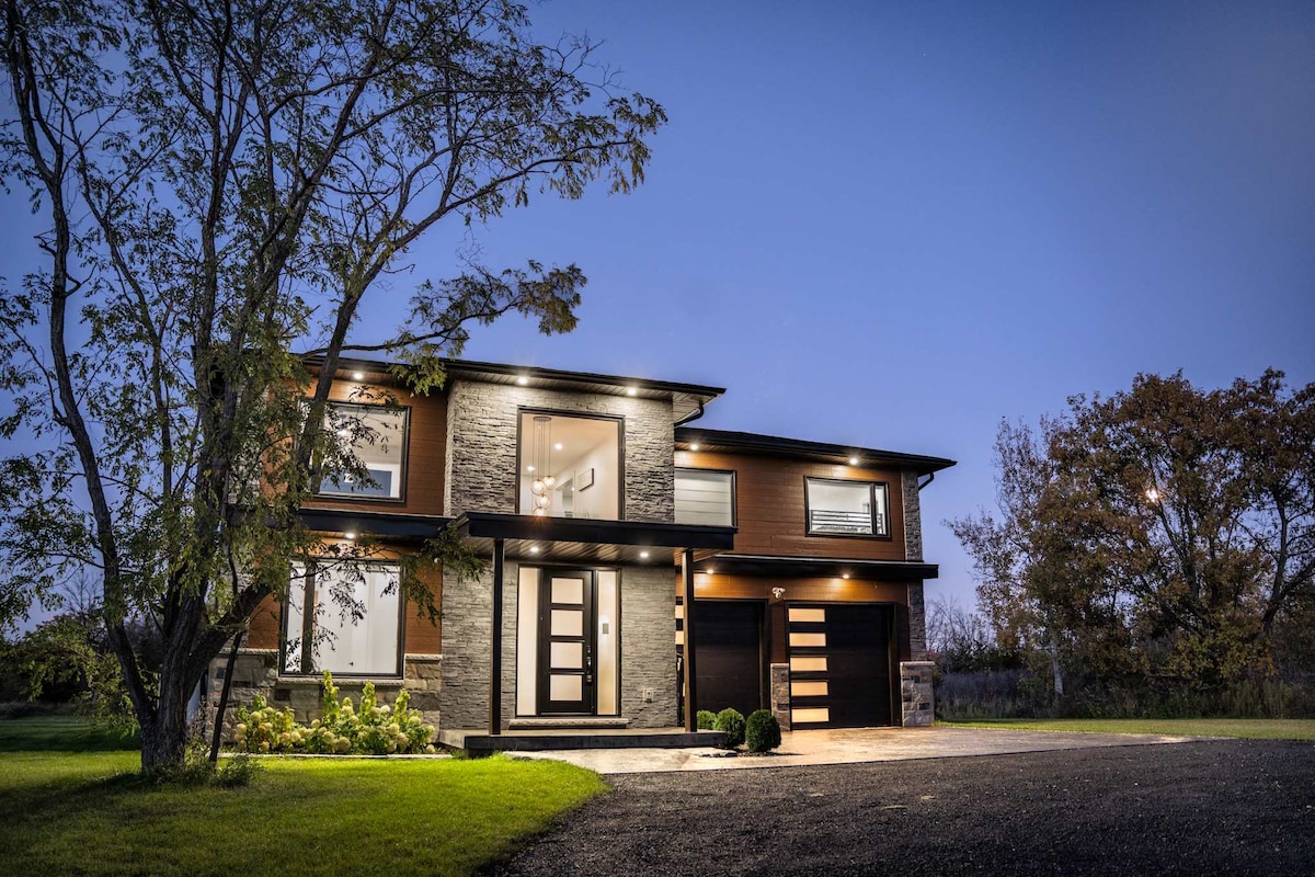 A modern two-story home showcases clean lines and a blend of natural stone and wood elements. Soft exterior lighting emphasizes the architectural features. Surrounding greenery adds a touch of nature, while a wide driveway leads to the garage entrance.