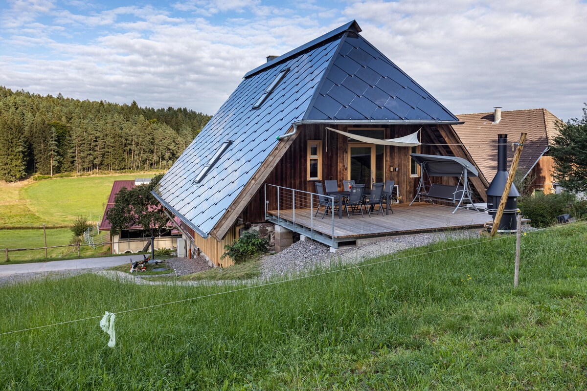A modern two-story building is set on a gentle slope, featuring a distinctive sloped roof with solar panels. An outdoor terrace is visible, furnished with a dining table and chairs. Lush green grass surrounds the property, and the background showcases a peaceful expanse of trees.