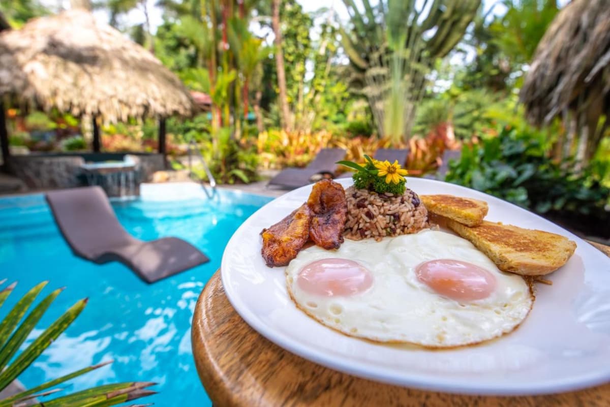 A plate of breakfast is presented with two fried eggs, rice and beans topped with herbs, crispy bacon, and golden plantain slices. The background features a refreshing outdoor pool surrounded by lush tropical foliage and a thatched roof seating area.