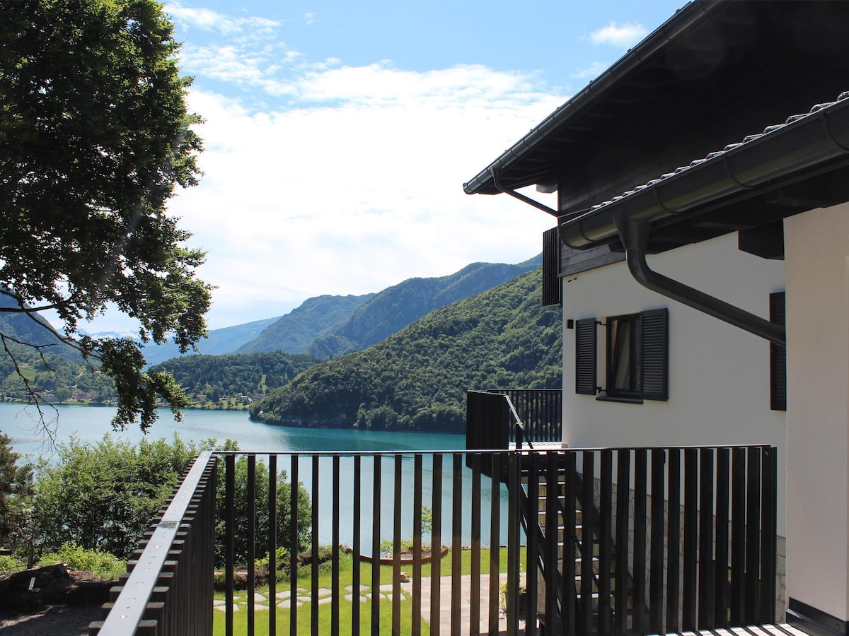 The exterior view features a modern villa adjacent to a tranquil lake, surrounded by lush mountains. A wooden balcony offers a vantage point for enjoying the scenic landscape and sunlight filtering through the trees, creating a peaceful outdoor setting.