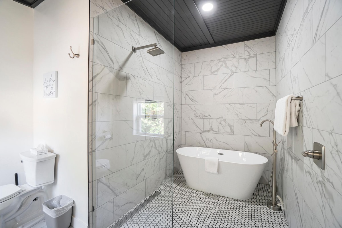 The modern bathroom features a spacious walk-in shower with sleek fixtures, alongside a freestanding bathtub. Light tiles with a subtle pattern cover the walls, while the floor is adorned with distinctive black and white hexagonal tiles. A window provides natural light, enhancing the serene atmosphere.