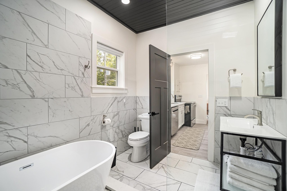 A spacious bathroom features elegant marble walls and flooring. A freestanding tub is positioned next to a glass-enclosed shower. A modern vanity with a mirror is visible, while a window allows natural light to brighten the space. Towels are neatly arranged for convenience.