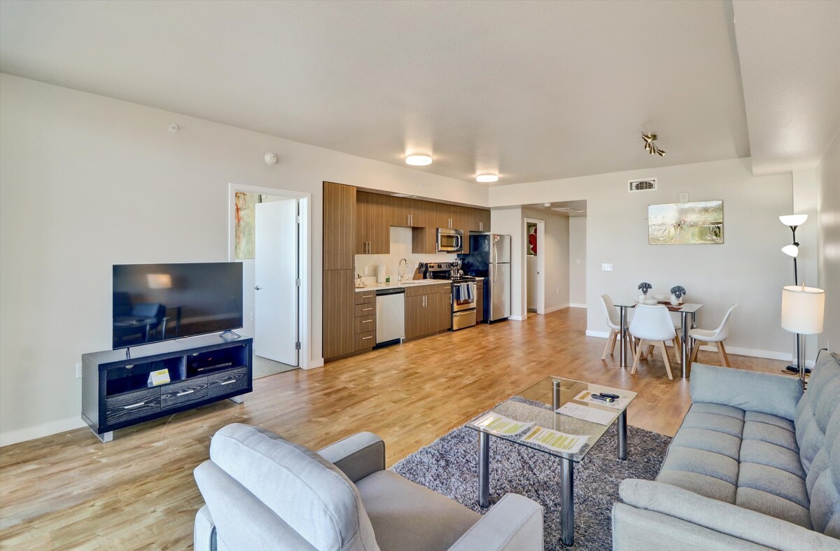 A modern living space is presented with a comfortable sofa and a sleek coffee table on a textured rug. In the background, a well-equipped kitchen features stainless steel appliances and cabinetry. A dining area with four chairs is visible, enhancing the open layout.