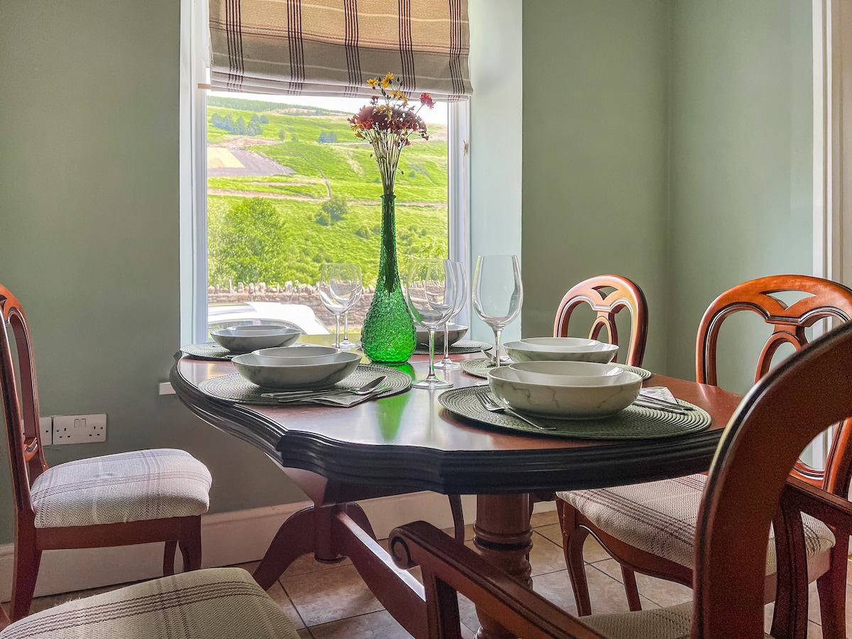 A dining table is set with plates and glassware, positioned near a window that offers views of the surrounding hills. A green vase with flowers adds a touch of color, complementing the soft tones of the walls and the comfortable wooden chairs.