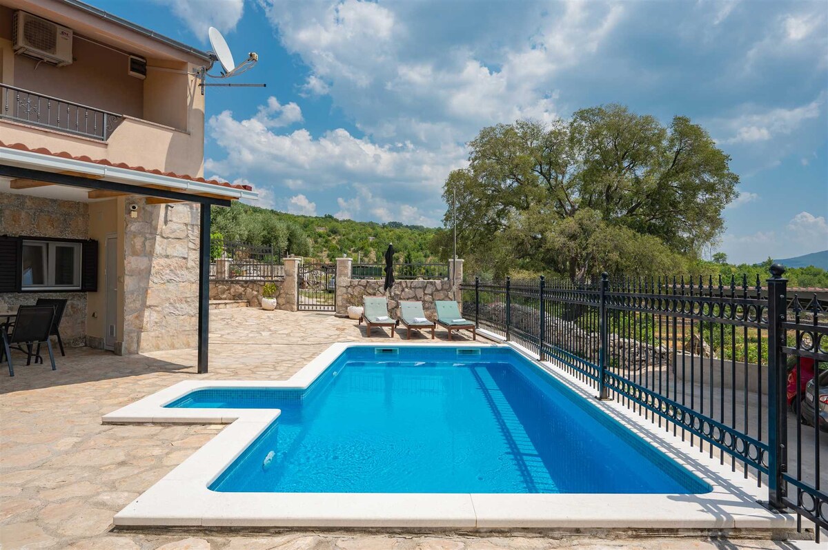 A private heated pool is set within the outdoor area, surrounded by a stone patio and five sun loungers. The landscape features lush greenery and a protective fence, creating a calm and secure environment. A shaded area for seating is visible nearby.