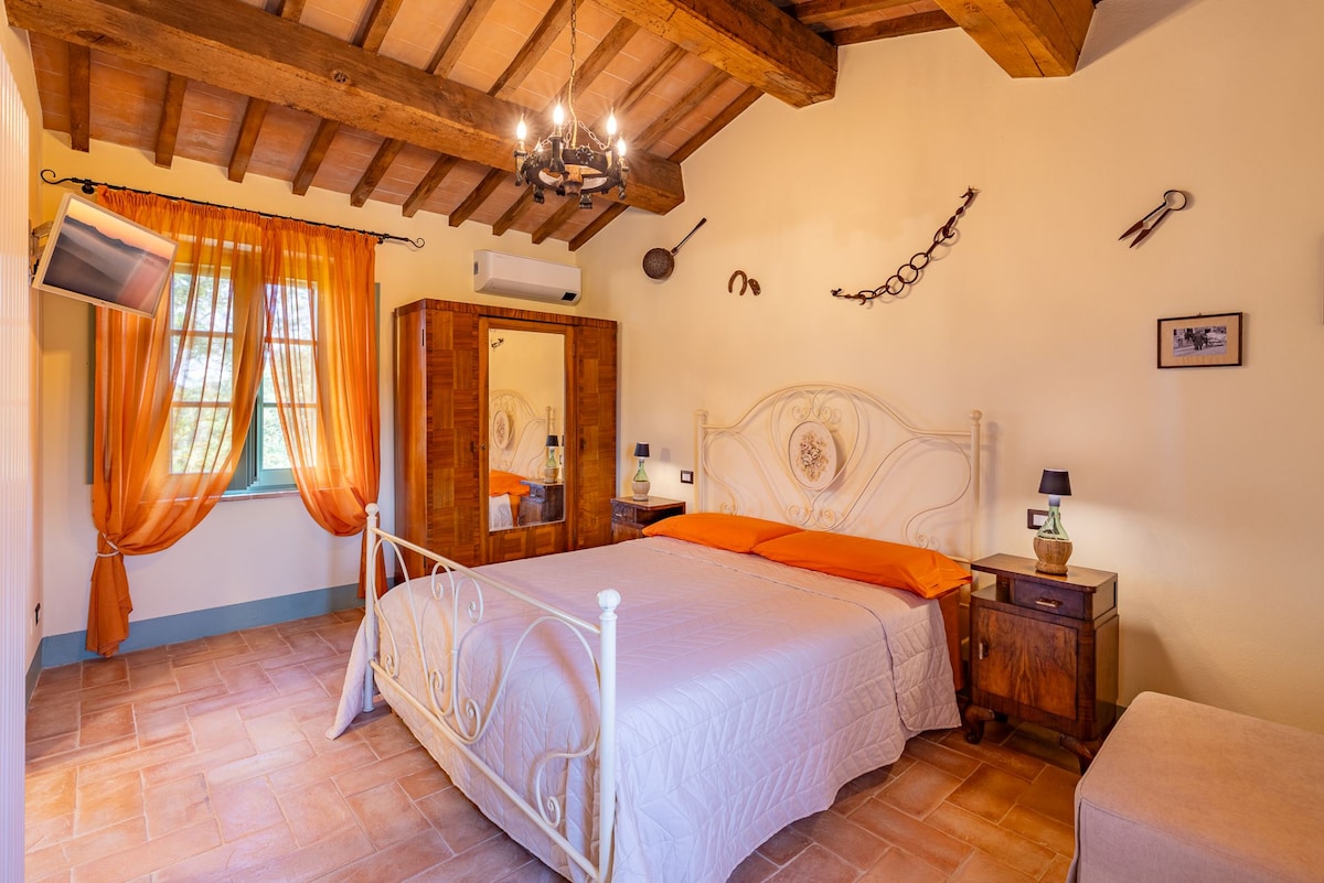 The room features a double bed with an orange accent, flanked by wooden bedside tables. A wardrobe reflects traditional Tuscan design, and a small TV is mounted on the wall. Light filters through orange drapes, enhancing the wood-beamed ceiling and terracotta floor.