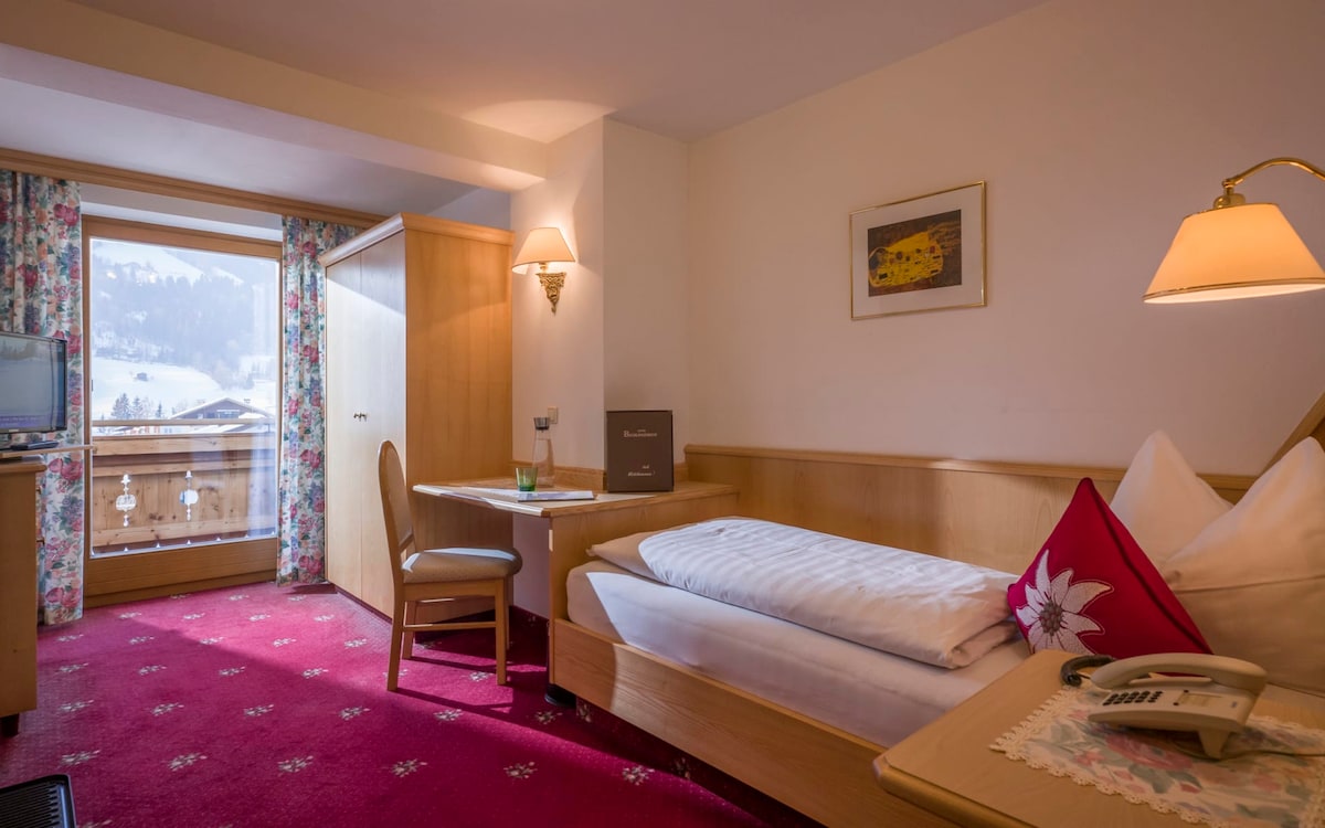 A cozy single room features a spacious bed with neutral bedding, a wooden desk, and a chair. Natural light enters through a balcony with mountain views. Soft lighting from a wall lamp enhances the warm wood accents in the room.