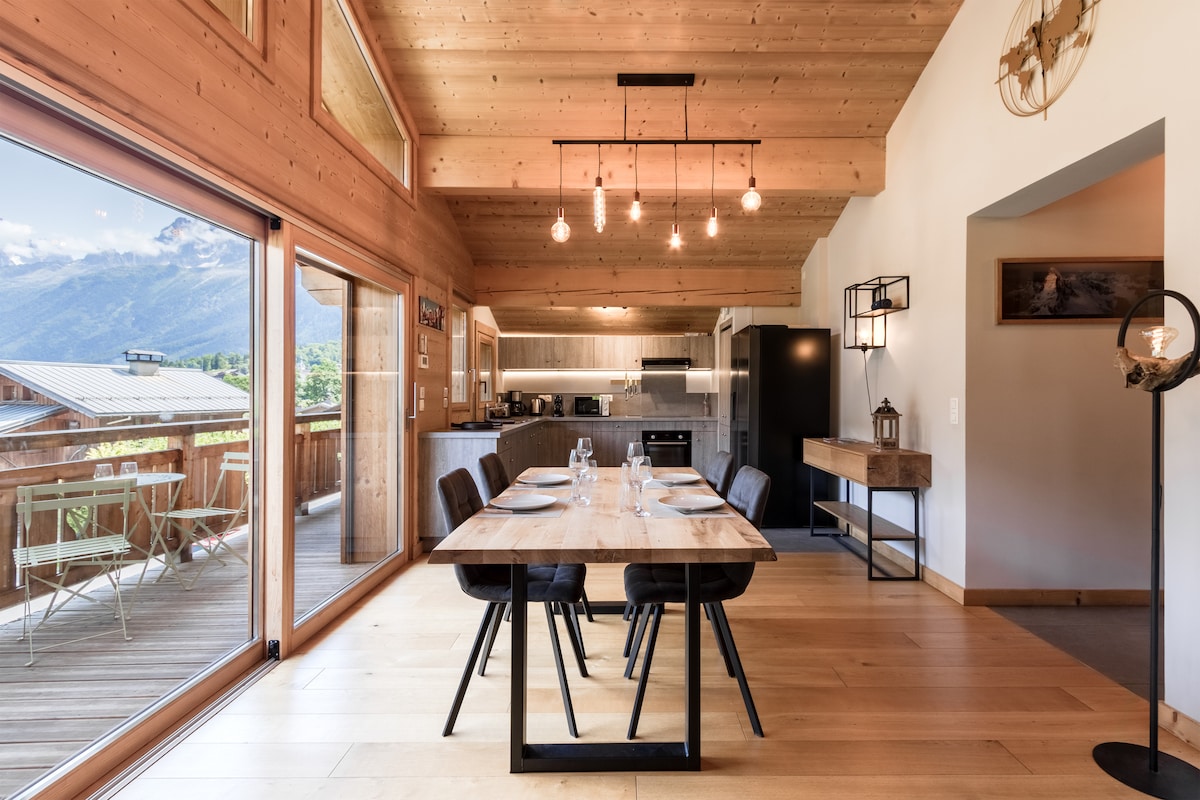 A spacious dining area features a wooden table set for four. The room is filled with natural light from large glass doors leading to a deck with mountain views. The ceiling showcases wooden beams, complementing the modern kitchen in the background.