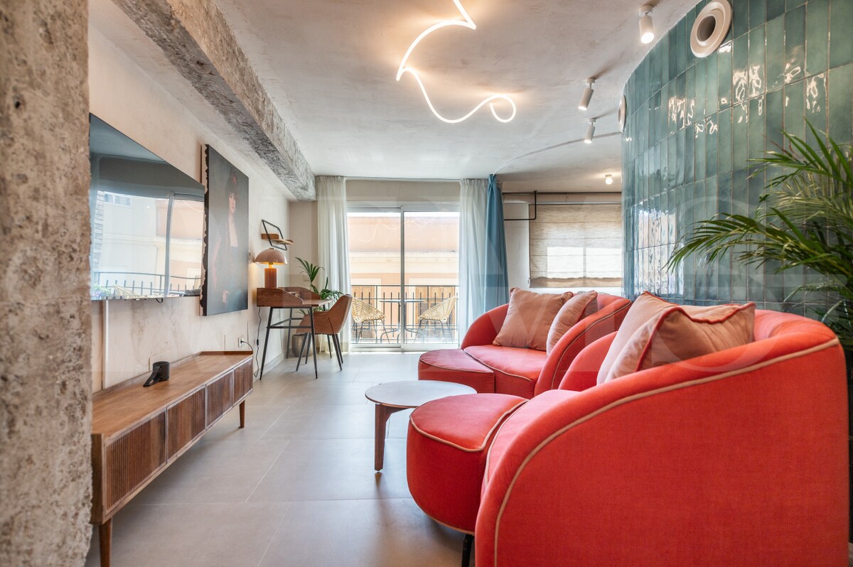 A spacious living area is furnished with two red sectional sofas, complemented by a low coffee table. A large wall-mounted television is positioned opposite the seating. Natural light filters in through glass doors leading to a balcony, while modern artwork adds a touch of character.