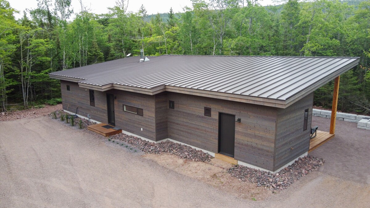 The exterior of the single-level home displays a modern design with a sloped metal roof. A combination of wood and stone elements creates a rustic feel. Surrounded by greenery, the entrance features a small deck with steps leading to the door.