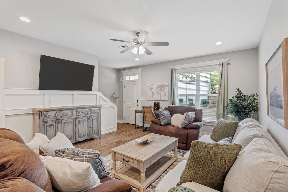A welcoming living room features comfortable seating arrangements, including a large sofa and armchairs, with a wooden coffee table centered in the space. A ceiling fan provides airflow, and large windows allow natural light to illuminate the room.