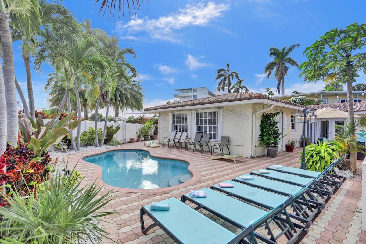 A tranquil outdoor area features a private heated pool surrounded by palm trees and tropical plants. Six chaise loungers are arranged neatly beside the pool. The patio is laid with pavers, leading to a charming façade of the house, with access to the entertainment spaces.