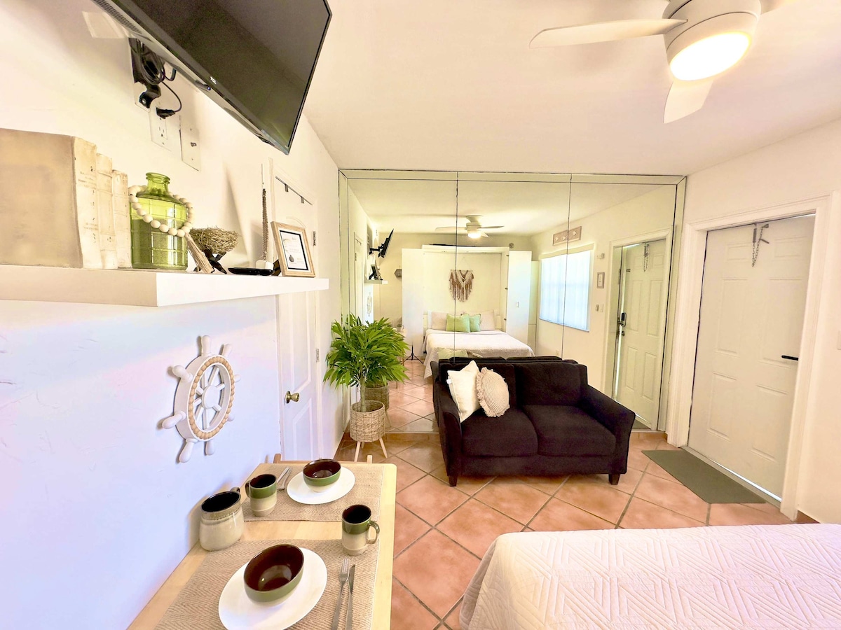 A cozy studio is highlighted by a dark sofa facing a dining table set with plates and bowls. A mirrored wall reflects the inviting sleeping area, which features a spacious bed. Light enters through a window, enhancing the airy feel of the space.