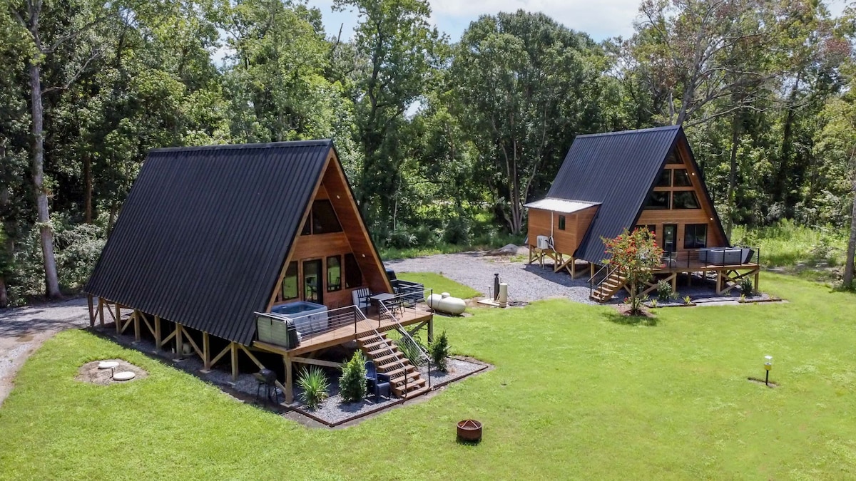 Two A-Frame cabins are situated in a lush green landscape, surrounded by trees. Each cabin features a sloped roof and large front decks. Hot tubs and seating areas are visible on the decks, creating inviting outdoor spaces.