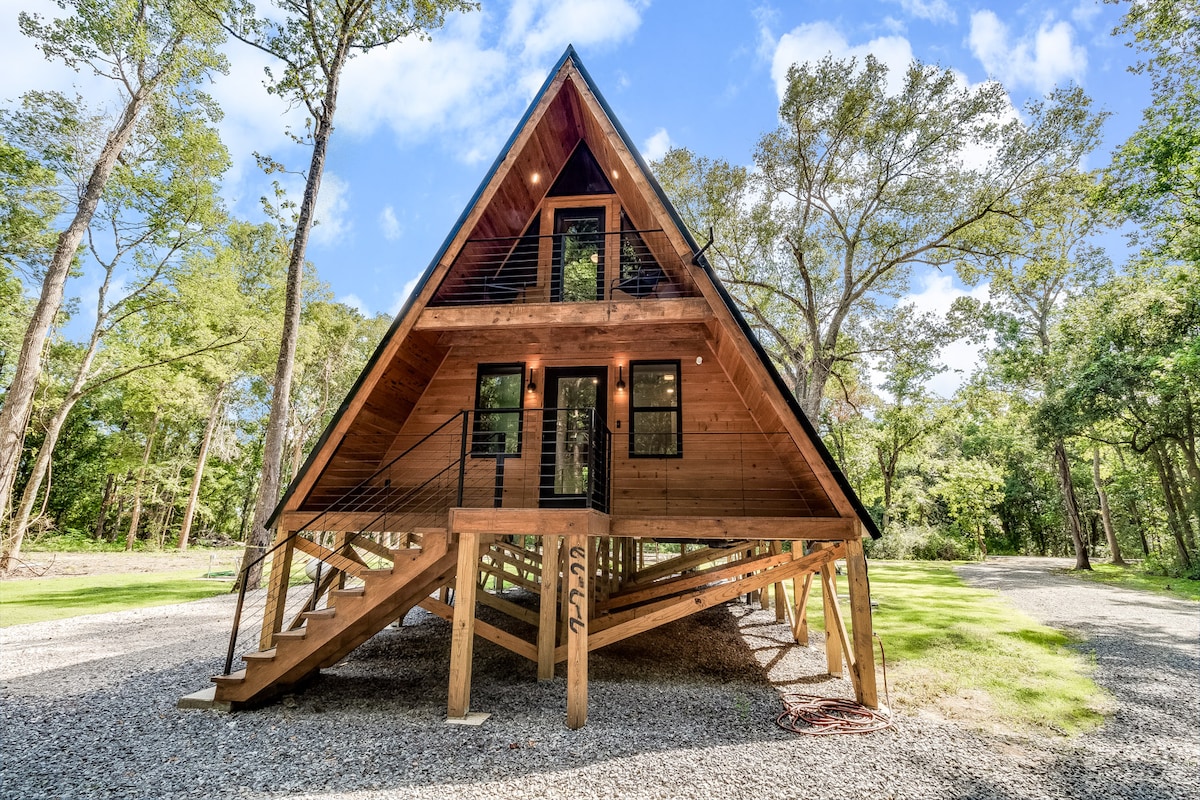The A-frame cabin features a striking triangular design with large windows on both levels. An inviting entrance is accentuated by a staircase leading up to the main door, surrounded by lush greenery and a gravel pathway, enhancing its natural setting.