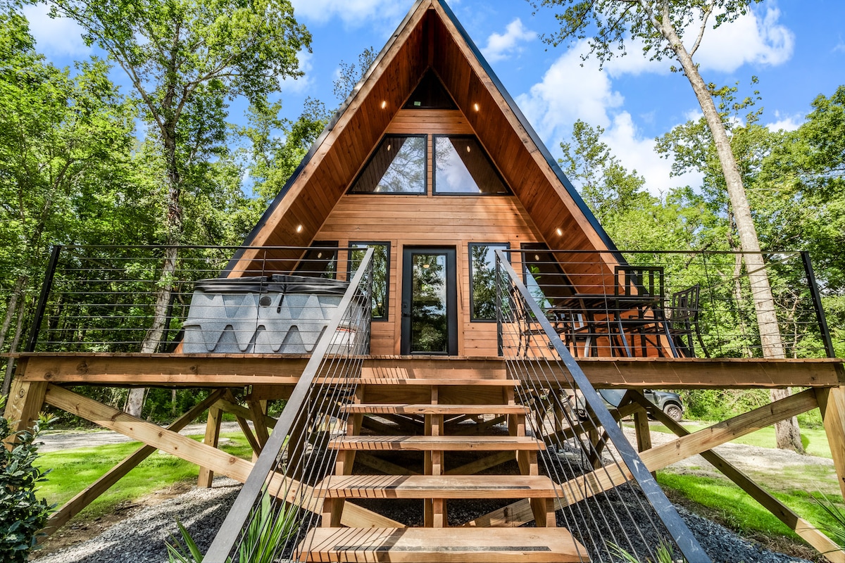 The A-Frame cabin is set on a raised platform, featuring a wooden exterior and a triangle-shaped roof. Large windows reach up to the peak, allowing ample natural light. A staircase leads to the entrance, surrounded by a lush, green landscape.