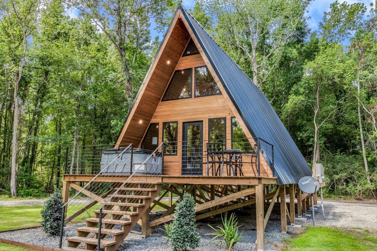A modern A-frame cabin is elevated above the ground, featuring a distinctive sloped roof and large front deck. The exterior includes wooden accents and expansive windows. Surrounding greenery contributes to a serene outdoor setting, while a hot tub rests on the deck.