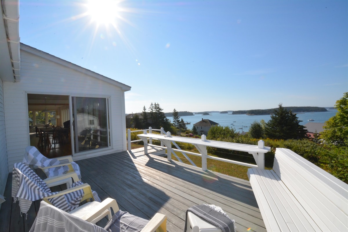An ocean view deck is bathed in sunlight, showcasing white outdoor furniture and soft, striped blankets. The panoramic vista includes the blue waters and distant shores dotted with homes and trees, inviting relaxation and enjoyment of the coastal scenery.